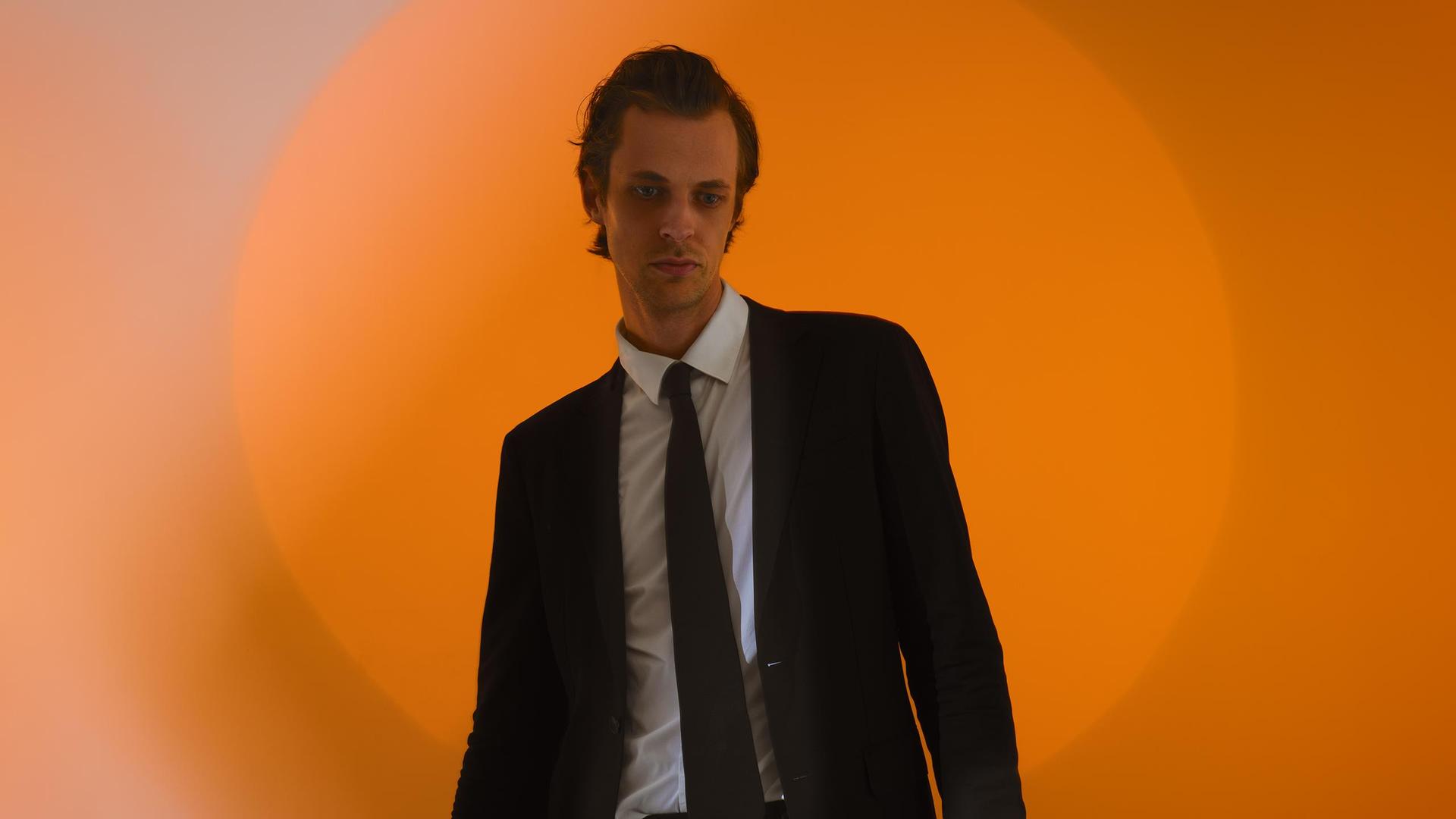 David Linszen, Promotiefoto van David Linszen in pak tegen een achtergrond met een oranje cirkel die vervaagd