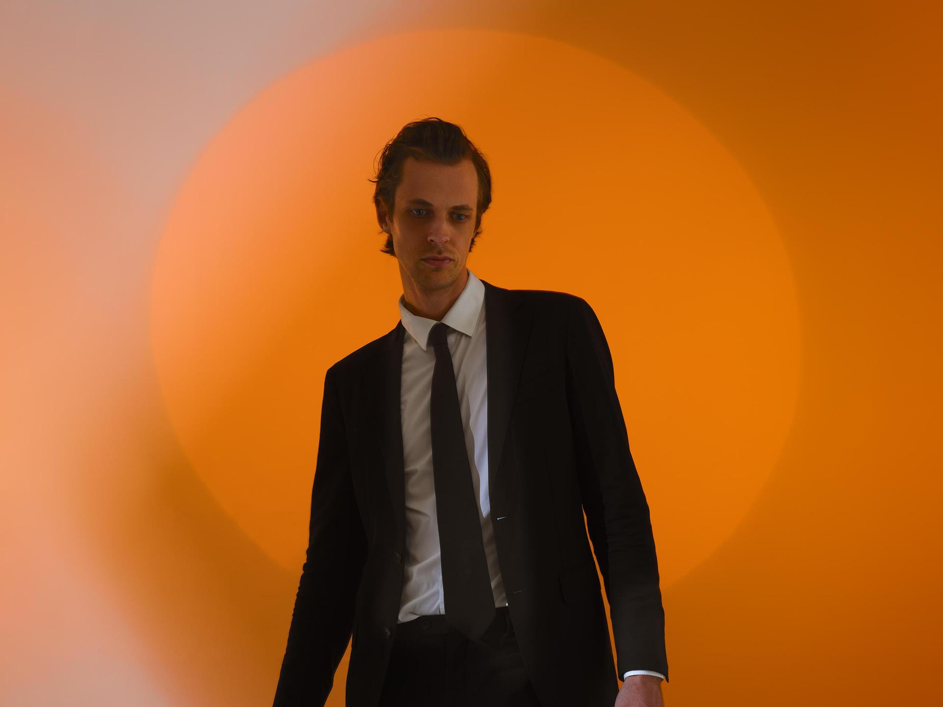 David Linszen, Promotiefoto van David Linszen in pak tegen een achtergrond met een oranje cirkel die vervaagd