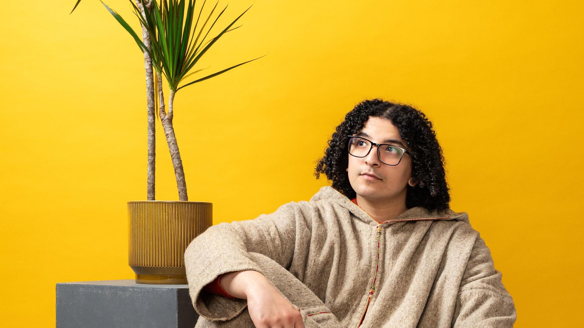 Ayoub, Promotiefoto van Ayoub zittend op de grond voor een gele wand met plant achter hem