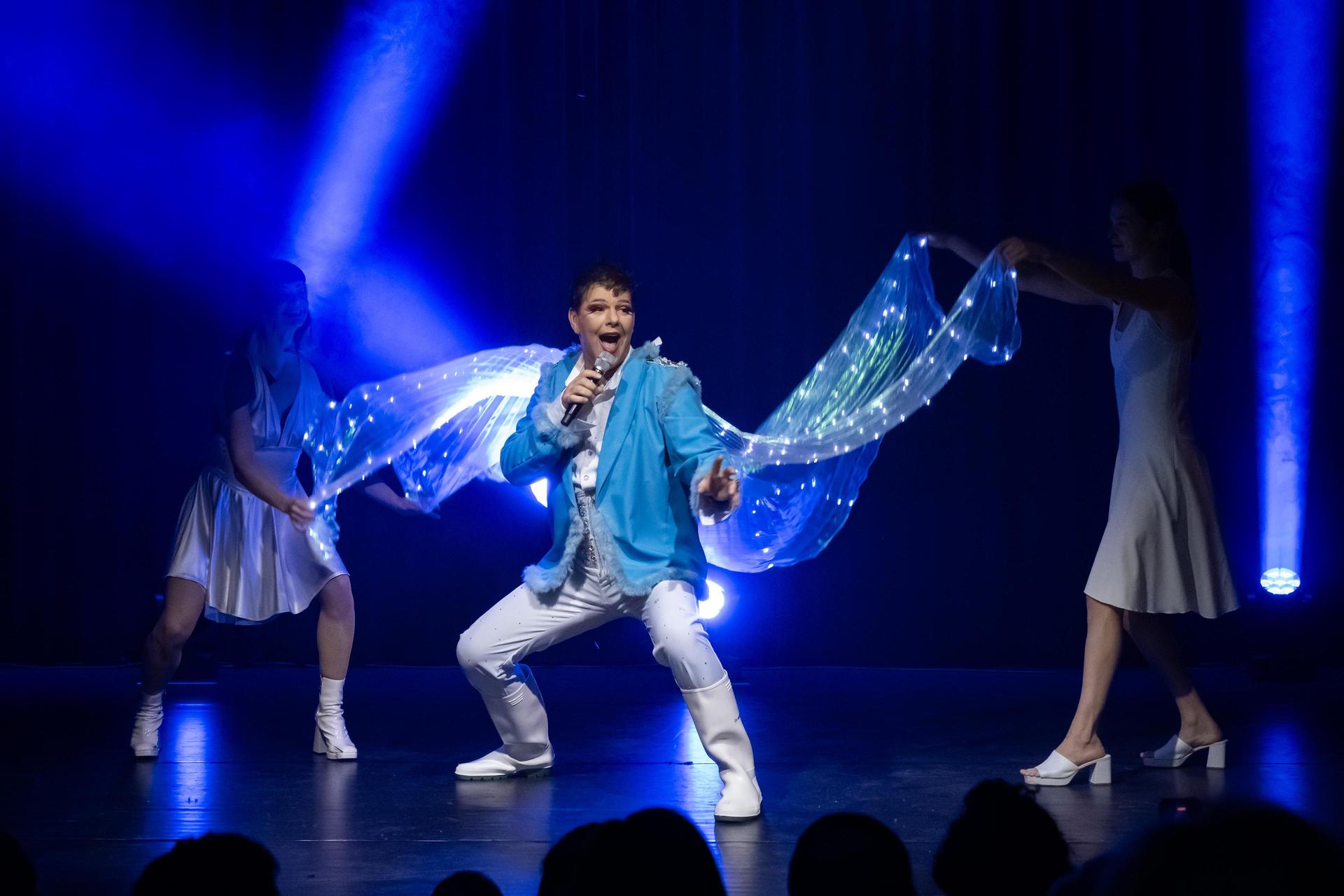 Foto Dave van der Wal zingend met een lichtblauw jasje aan met slepen die licht geven en zweven