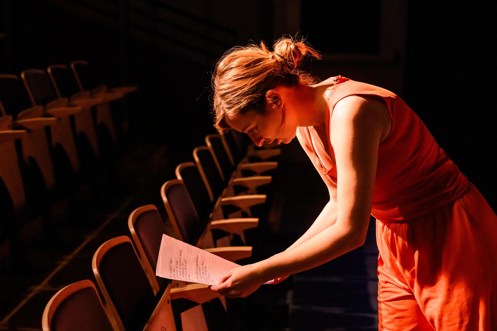 scènefoto met Anne Neuteboom in oranje outfit bij opgeklapte theaterstoelen