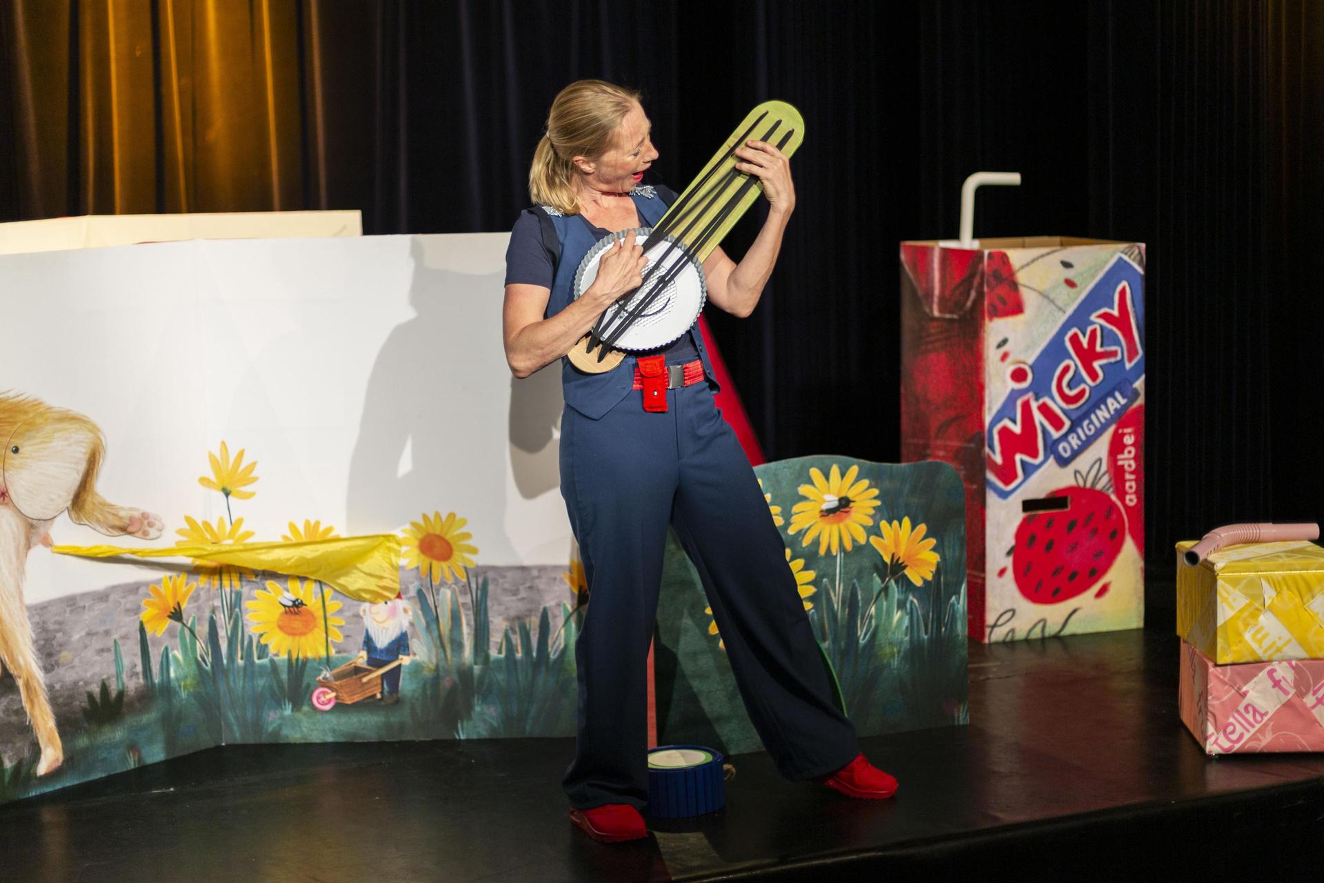 scènefoto Het Kabouterboek met actrice spelend op soort gitaar in decor met oa grote bloemen