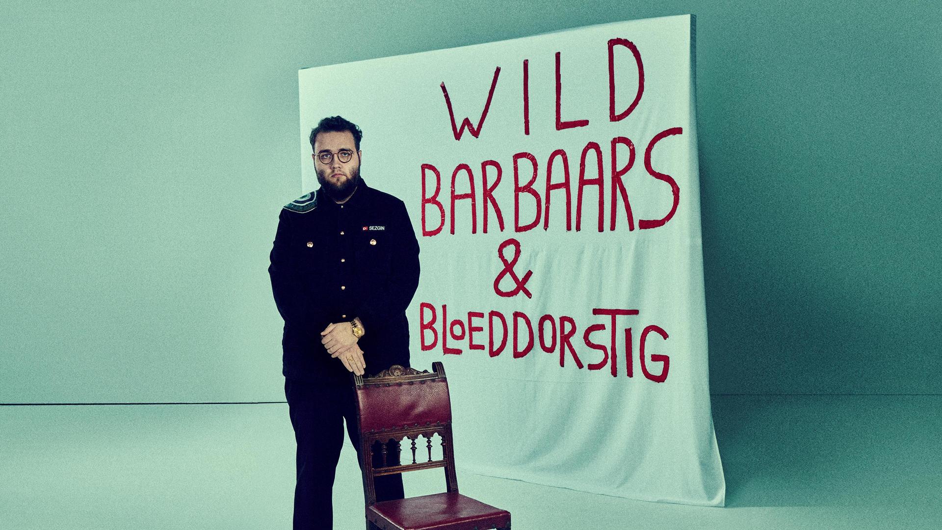 Promotiefoto van Sezgin Gulec met een doek achter hem met de titel Wild, Barbaars en Bloeddorstig