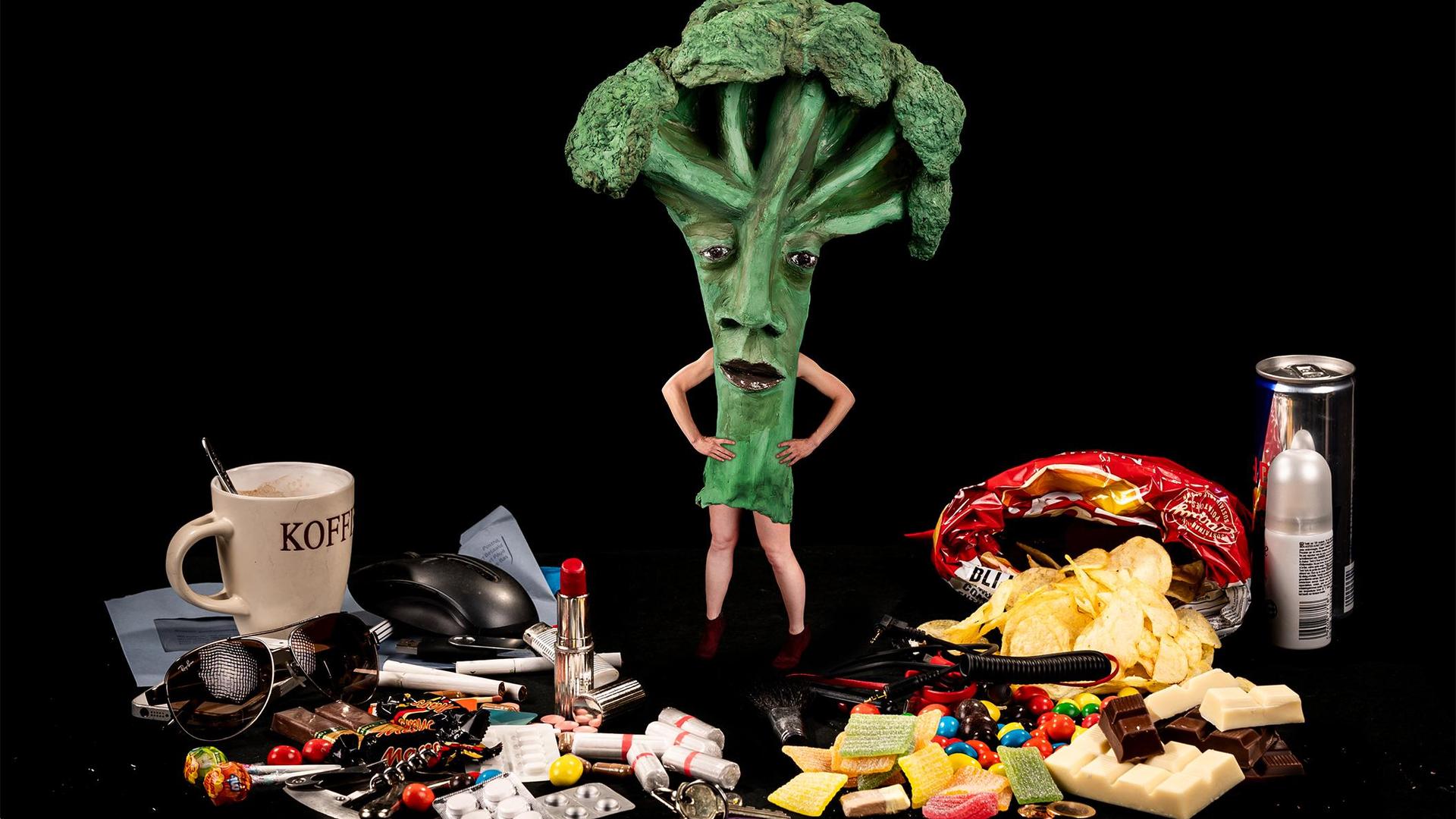 Foto met vrouw verkleed als broccoli op tafel met heel veel grote mensen spullen: medicijnen, koffie