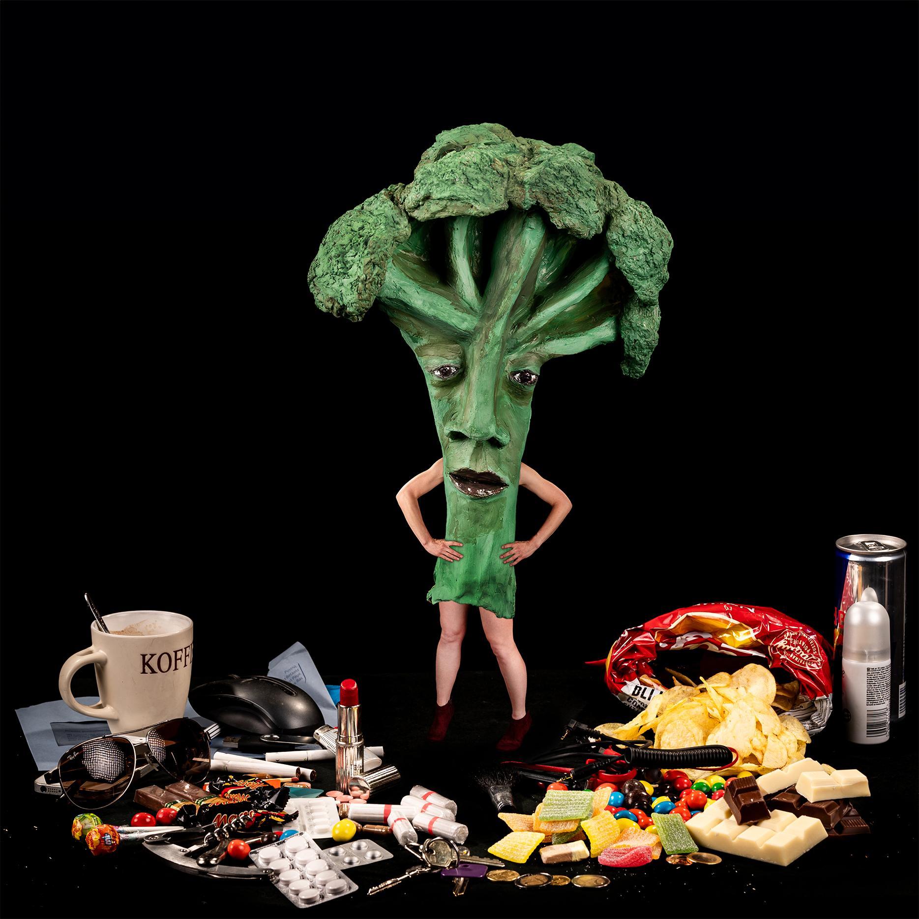 Foto met vrouw verkleed als broccoli op tafel met heel veel grote mensen spullen: medicijnen, koffie