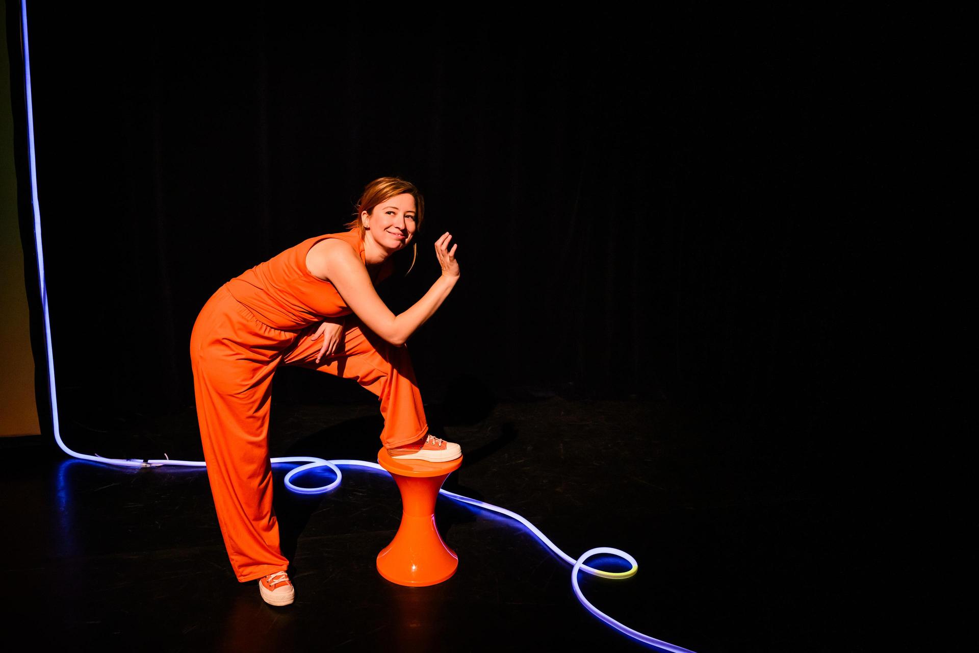 scènefoto met Anne Neuteboom in oranje outfit tegen zwarte achtergrond