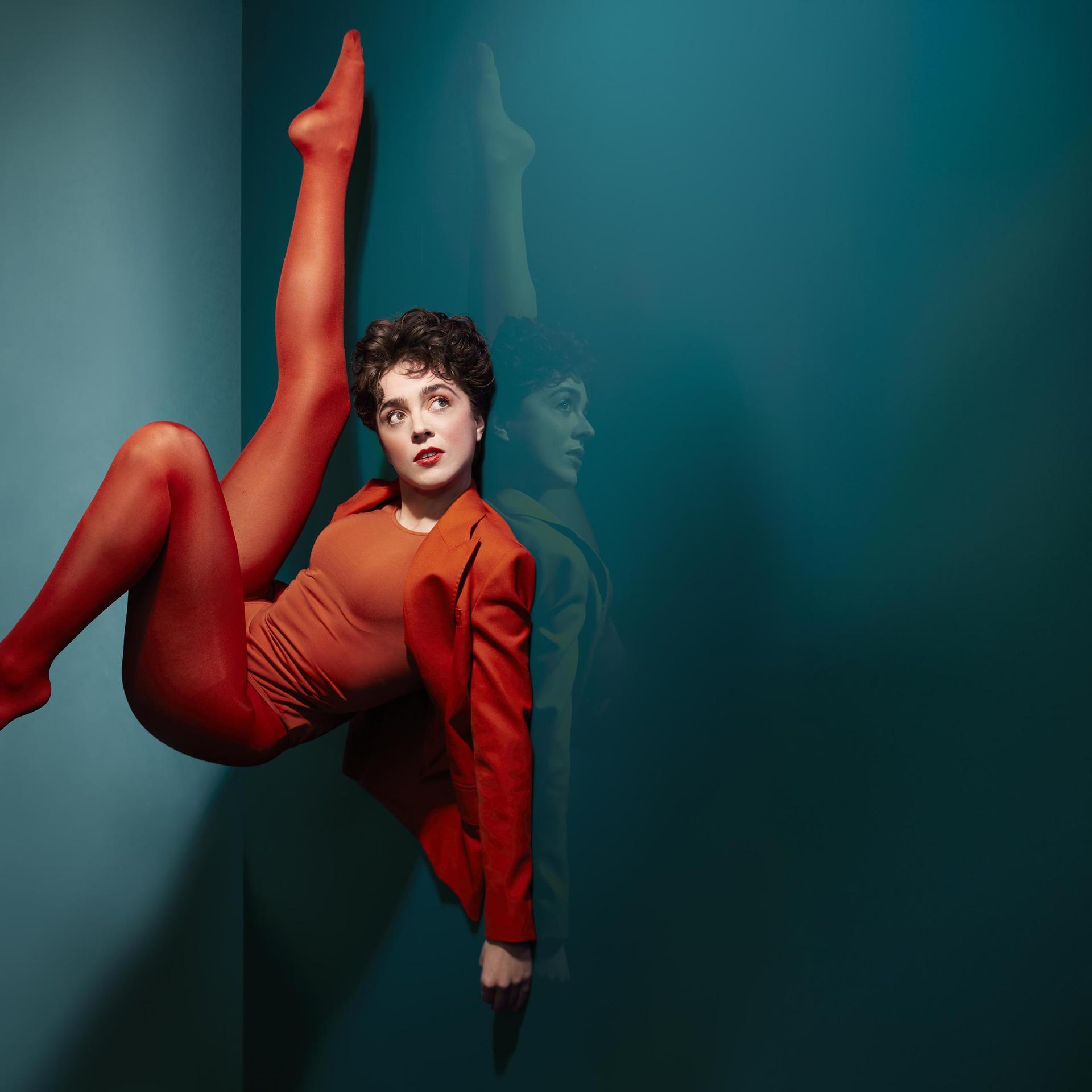Selma Visscher, Promotiefoto Sema Visscher in rood pak tegen groene wand met haar benen in de lucht